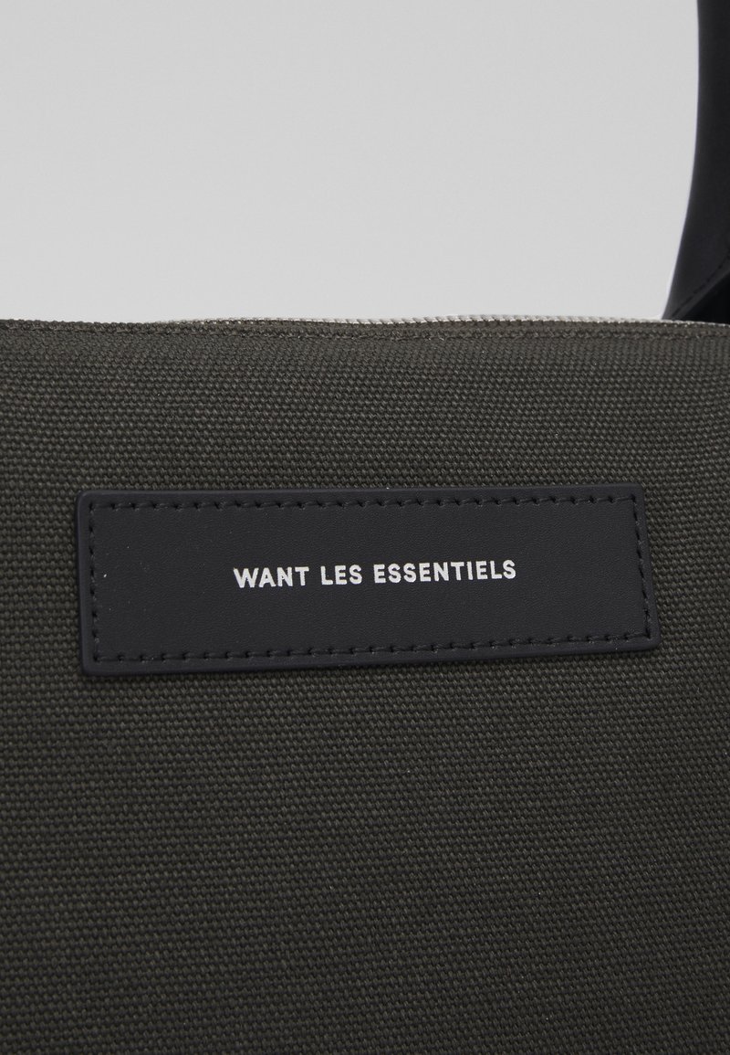 Dunkelgrüne Stofftasche mit einem schwarzen Lederanhänger, auf dem der eingestempelte weiße Text "WANT LES ESSENTIELS" steht. Strukturierte Leinenaußenfläche.