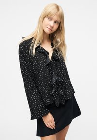 Νεαρή γυναίκα με μακριά ξανθά μαλλιά που φοράει μαύρο πουκάμισο με πουά και βολάν και μαύρη φούστα, σε απλό λευκό φόντο.