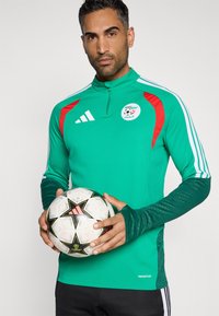 Zöld sportos hosszú ujjú felső, piros díszítéssel, fehér csíkokkal és futball emblémával, egy mintás fekete-fehér focilabdával párosítva.