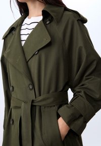 Oliven grøn trenchcoat med dobbeltknaplukning, oversized krave, bælte i taljen og knapdetaljer. Lavet af glat stof.