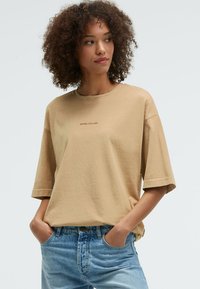 Beige oversized t-shirt with short sleeves, round neckline, and center text "SENSES.THE LABEL." Paired with light blue denim jeans.