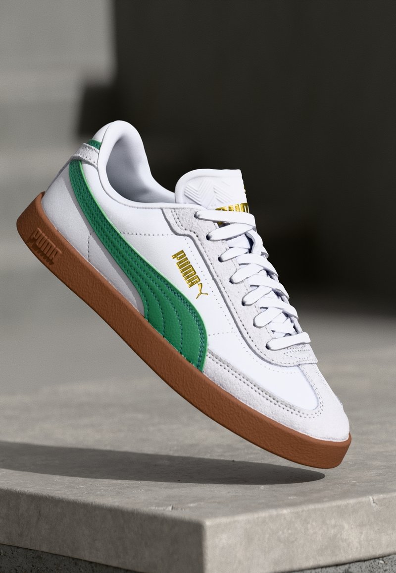 Sneaker bianco con dettagli verdi e logo dorato. Presenta una suola in gomma testurizzata, un design tradizionale con lacci e un colletto imbottito per il comfort.