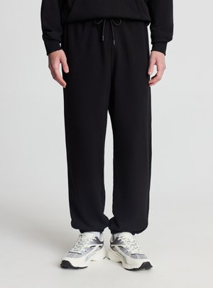 Parte bassa del corpo di una persona che indossa pantaloni della tuta neri con coulisse in vita e sneaker sportive bianche e grigie su uno sfondo chiaro e semplice.