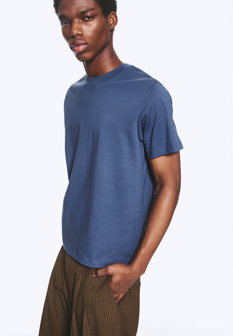 Jeune homme portant un t-shirt bleu à manches courtes et un pantalon marron à fines rayures, une main dans la poche, sur un fond blanc uni.