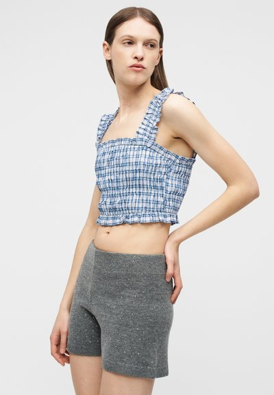 Jeune femme portant un crop top volanté à carreaux bleus et blancs et un short en maille gris avec de petites décorations, posant devant un fond uni.