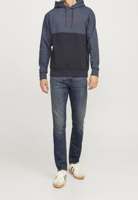 Sudadera azul marino con una sección superior de un azul más claro, cordones y texto rojo; combinada con jeans de mezclilla y zapatillas blancas con detalles verdes.
