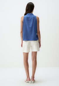 Blaue ärmellose Bluse mit Kragen, aus leichtem Stoff, kombiniert mit weißen Shorts. Glatte Textur mit lockerer Passform und seitlichen Schlitzen.