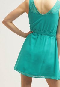 Femme portant une robe sans manches couleur sarcelle avec des détails en dentelle sur le haut du dos, se tenant avec une main sur la hanche.