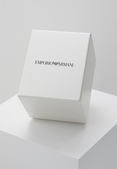Scatola rettangolare bianca con il logo Emporio Armani nero, posizionata inclinata su un piedistallo bianco su sfondo grigio chiaro.