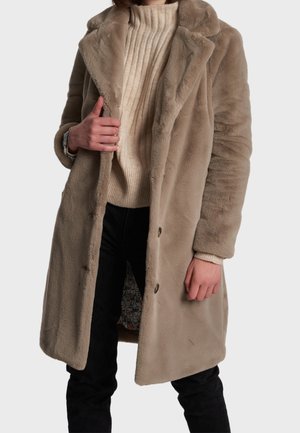 Personne portant un manteau en fausse fourrure beige par-dessus un pull côtelé crème et un pantalon noir, tenant le manteau ouvert pour révéler une doublure à motifs.