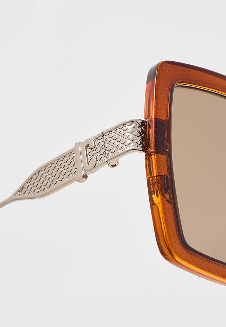 Lunettes de soleil surdimensionnées amber avec des verres marron, dotées d'une charnière en métal argenté texturé et d'un design de monture angulaire.