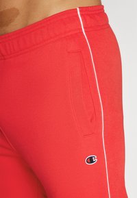 Pantalones de chándal rojos hechos de una tela suave, con una franja lateral blanca y un parche de logo en el muslo. Incluye bolsillos en cada lado.