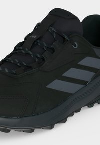 Chaussure de sport noire avec semelle texturée, trois bandes grises sur le côté, col rembourré et lacets à motif passés à travers des boucles en tissu.