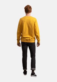 Felpa gialla con polsini e orli a coste, abbinata a jeans neri arrotolati alle caviglie. Modello di spalle, che indossa sneaker nere.