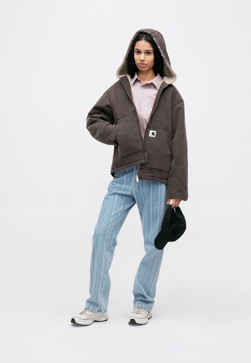 Brown hooded jacket with a fleece lining, large pockets, and a logo patch, paired with striped light blue jeans and sneakers.