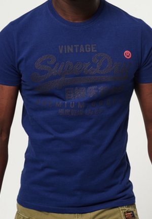 Homme portant un t-shirt bleu ajusté « Vintage SuperDry Premium Goods » et un pantalon beige sur un fond uni.