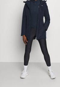 Niebieska wodoodporna kurtka z kapturem, zamek z przodu; czarne teksturowane legginsy; białe sneakersy z sznurowadłami i teksturowanymi podeszwami, widoczne białe skarpetki do kostek.