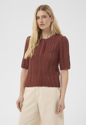 Frau mit langen blonden Haaren, trägt ein kurzärmliges, strukturiertes braunes Strickoberteil und cremefarbene Hosen und steht vor einem schlichten, hellen Hintergrund.