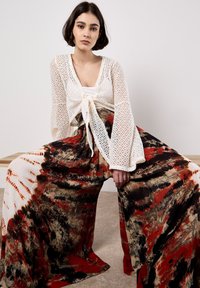 Haut en dentelle blanche à manches longues avec un nœud sur le devant, associé à un pantalon à larges jambes à motifs dans des teintes rouges, brunes et noires. Texture lisse.
