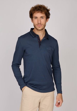 Homme aux cheveux bouclés portant un polo à manches longues bleu marine et un pantalon beige, se tenant avec une main dans la poche devant un fond uni.