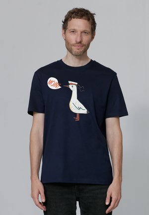 Marineblauwe katoenen t-shirt met een witte geïllustreerde vogel met rode accenten en een spraakballon met de tekst "MOIN!" op de voorkant. Korte mouwen, ronde hals.