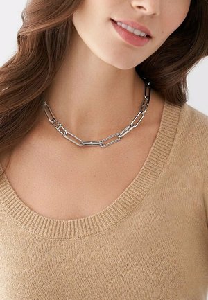 Silberne Metallkettenschmuck mit verlängerten Gliedern und polierter Oberfläche, getragen über dem Ausschnitt eines beigen, gerippten Pullovers.