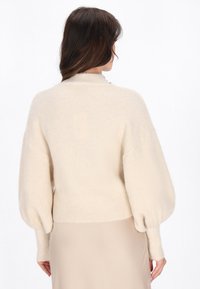 Crème-kleurige trui met een zachte textuur, hoge halslijn en ballonmouwen. Aansluitend bij de taille, gecombineerd met een beige rok.