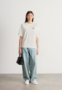 Biele oversize tričko s čiernym logom na hrudi, v kombinácii so svetlomodrými voľnými džínsami a bielymi teniskami, držiace malú čiernu kabelku.