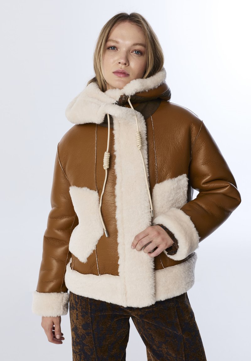 Bruine leren jas met crèmekleurige fleece accenten, een capuchon en trekkoorddetails. Heeft een boxy silhouet en textuurpatronen op de stof.