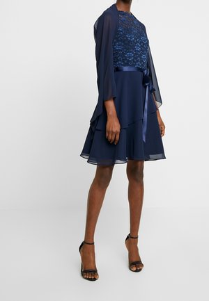 Frau trägt ein knielanges marineblaues Kleid mit Spitzenoberteil, transparenten, mehrlagigen Ärmeln und Satinband-Gürtel, kombiniert mit schwarzen Schuhen mit Knöchelriemen.