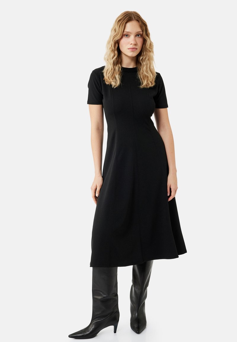 Femme aux cheveux blonds ondulés portant une robe midi noire à manches courtes et des bottes noires en cuir montantes jusqu'aux genoux, debout devant un fond clair uni.