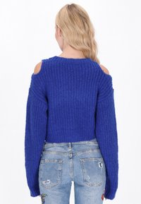 Maglione blu lavorato a maglia con design a spalle scoperte, texture a coste e maniche oversize, abbinato a jeans in denim chiaro strappati.