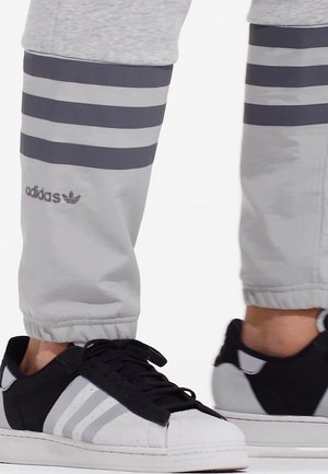 Nærbillede af underben iført lysegrå Adidas bukser med tre sorte striber, parret med sort-hvide Adidas sneakers.