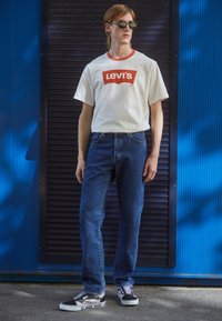 Ung man i vit Levi's t-shirt, blå jeans, Vans sneakers och solglasögon som står mot en blå och svart strukturerad vägg utomhus.