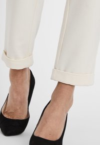 Pantaloni su misura color crema con risvolti, abbinati a scarpe con tacco alto in suede nero. Il tessuto ha una texture liscia.