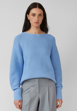 Frau mit dunklen Haaren, die einen strukturierten, hellblauen Strickpullover und graue, gestreifte Hose trägt, auf einem einfachen Hintergrund stehend.