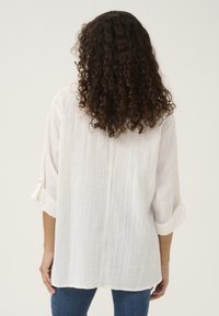 White, textured oversized shirt with rolled sleeves and button accents. View from the back, paired with denim jeans.