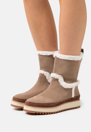 Brown suede boots with white faux fur trim, a rounded toe, and a textured sole. The height reaches mid-calf, featuring contrast stitching.