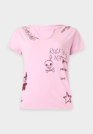 Γυναίκα με ροζ γραφικό t-shirt και μακριά καφέ φούστα με πλευρική σκίσιμο, στέκεται μπροστά σε απλό φωτεινό φόντο.