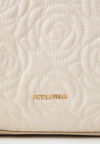 Sac à main beige matelassé avec des motifs de roses embossés et un logo doré "ANNA FIELD". Texture lisse avec une forme structurée.