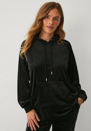 Kobieta o długich włosach, ubrana w czarną welurową bluzę z kapturem, kieszenią z przodu i sznurkami, pozująca na tle jednolitego jasnego tła.