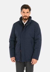 Giacca isolante blu navy con colletto alto, cerniera frontale, bottoni a pressione e tasche laterali, caratterizzata da una texture liscia e opaca.