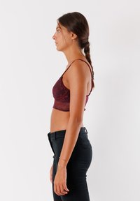 Donna con i capelli intrecciati, indossa un crop top in pizzo bordeaux e pantaloni neri, in posizione di profilo contro uno sfondo bianco semplice.