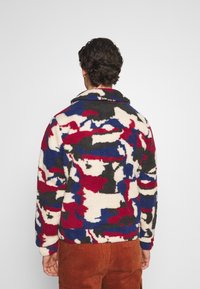 Fleece-jacka med ett flerfärgat abstrakt kamouflage-mönster i röd, blå, svart och kräm. Har krage och texturerad tyg.