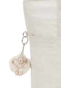 Beige textiel tas met een pluizige crèmekleurige sleutelhanger in de vorm van een gestileerde aap, met een metalen clip bevestigd aan de zijzak.