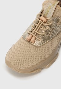 Beige sneakers med strikket mesh-overdel, elastiske lisser med toggle og glitrende gull-detaljer på en lys såle.