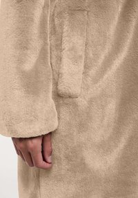 Beżowy płaszcz z sztucznego futra o fakturowanej powierzchni, z długimi rękawami i boczną kieszenią. Materiał wydaje się miękki i puszysty.