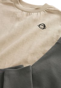 Hellbeiges Baumwoll-T-Shirt mit geripptem Rundhalsausschnitt, versehen mit einem schwarzen Affenlogo auf der Brust und grauen, gerippten Ärmeln.