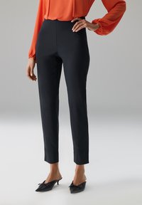 Zwarte op maat gemaakte broek met een strak ontwerp, hoge taille en een gladde textuur, gecombineerd met puntige zwarte mules met een decoratieve strik.