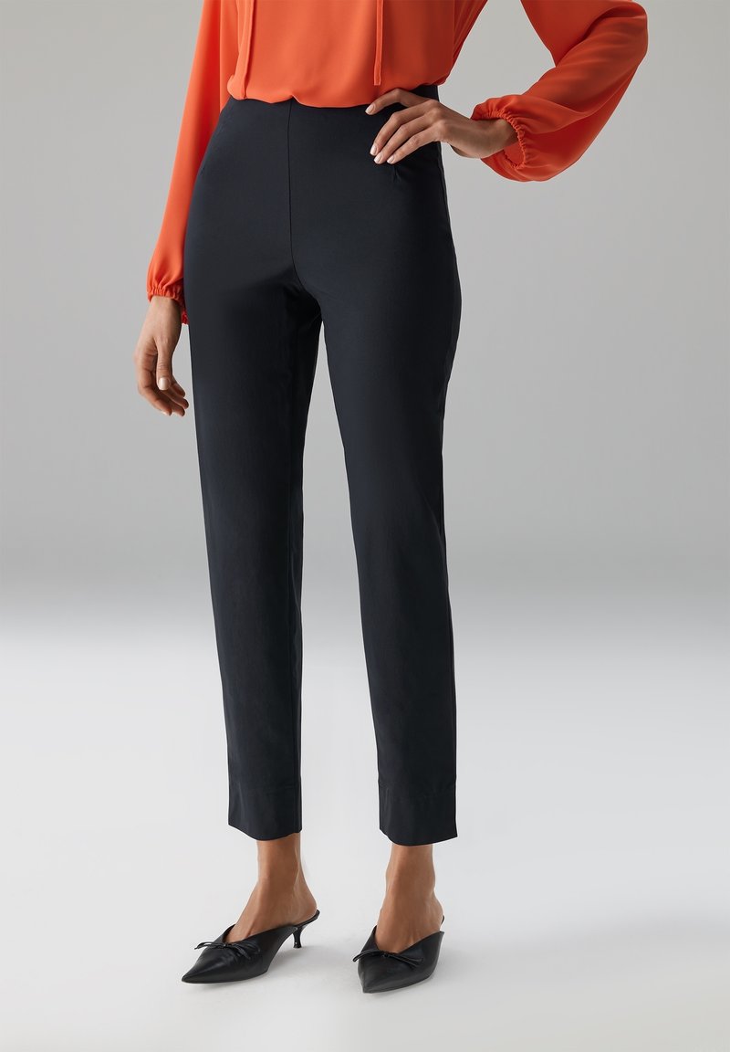 Zwarte op maat gemaakte broek met een strak ontwerp, hoge taille en een gladde textuur, gecombineerd met puntige zwarte mules met een decoratieve strik.